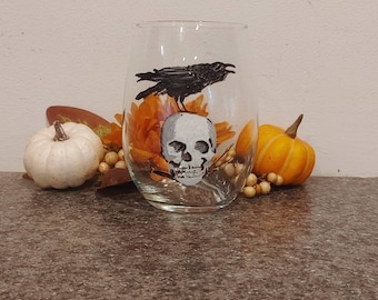 May include: A clear stemless wine glass with a hand-painted Halloween design. The design features a white skull with a black crow perched above it. The glass is surrounded by orange flowers and small pumpkins.