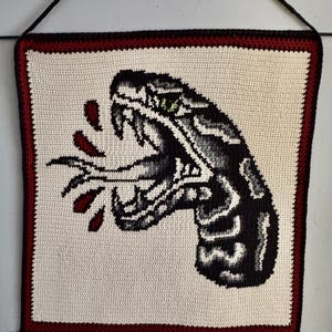 May include: Crocheted wall hanging depicting a black and grey snake head with an open mouth, fangs, and green eyes. Red blood droplets are shown. The artwork is framed with a red and black border on a cream background.