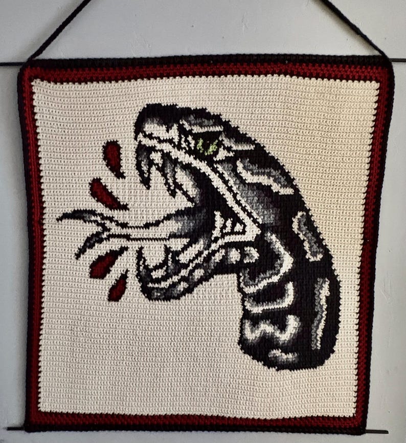 May include: Crocheted wall hanging depicting a black and grey snake head with an open mouth, fangs, and green eyes. Red blood droplets are shown. The artwork is framed with a red and black border on a cream background.