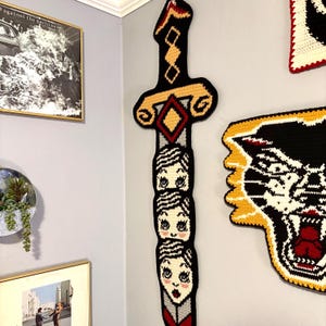 May include: A large, black, yellow, and red crocheted sword-shaped wall hanging. The sword features a decorative handle and a blade adorned with three stylized faces. The wall hanging is a unique piece of home decor.