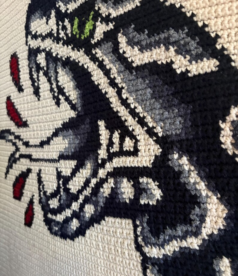 May include: Close-up of a cream-coloured crocheted textile featuring a detailed design of a snake's head. The snake is depicted in shades of black, grey, and white, with red accents and a green eye. The crochet stitches are visible.