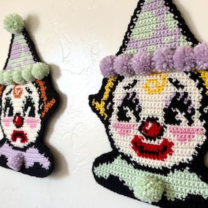 May include: Two crocheted clown wall hangings in different sizes. Each clown features a pointed hat with a pom-pom, a white face with black eye details, a red nose, pink cheeks, and a ruffled collar. The larger clown has a red mouth, while the smaller clown has a downturned mouth. Both are outlined in black yarn.