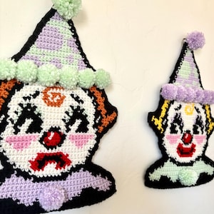 May include: Two crocheted clown wall hangings featuring white faces, red noses, and black outlines. The larger clown wears a lavender and mint green hat with mint pom-poms, while the smaller clown has a lavender hat and a mint green collar.