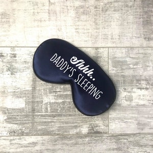 May include: A navy blue sleep mask with the words "Shhh.. DADDY'S SLEEPING" in white. The mask is made of a smooth, satin-like material. The sleep mask is on a light grey wooden surface.