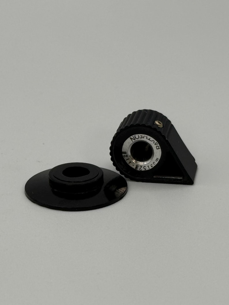 May include: Two black plastic components. One is a circular base with a central hole, and the other is a triangular piece with a ridged top and a circular inset with text. The text reads "DAVTICON" and "82515-1128".