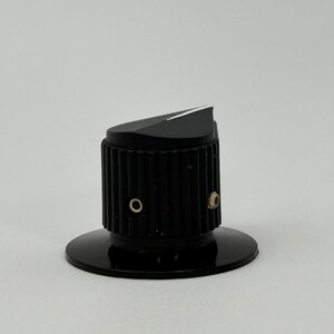 May include: A black control knob with a ridged cylindrical body and a slanted top. The knob has a circular base and two small holes. The top surface is angled with a white accent.
