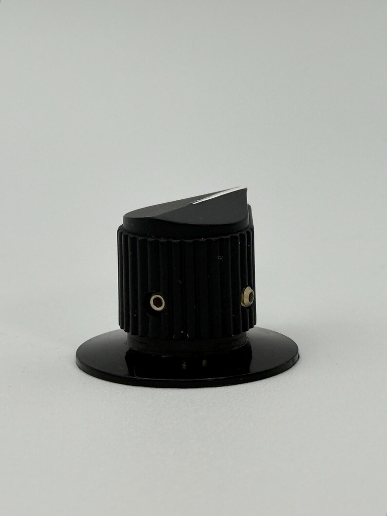 May include: A black control knob with a ridged cylindrical body and a slanted top. The knob has a circular base and two small holes. The top surface is angled with a white accent.