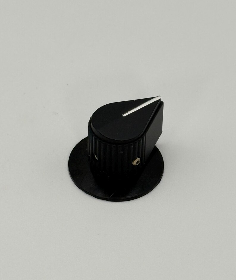 May include: A black control knob with a white indicator line. The knob has a ridged cylindrical base and a triangular top. It is mounted on a circular black base, suitable for electronic devices.