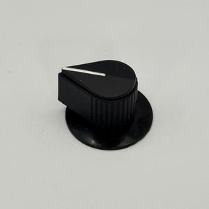May include: A black control knob with a white indicator line. The knob has a rounded top and a ridged base, sitting on a circular base. The image is a close-up shot against a plain white background. This component is likely for electronics or audio equipment.