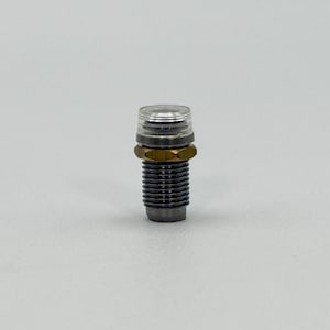 May include: A cylindrical metal component featuring a threaded black body, a central brass-colored hexagonal nut, and a clear, translucent plastic cap at the top. The object is designed with a ridged exterior and a flat, circular top surface.