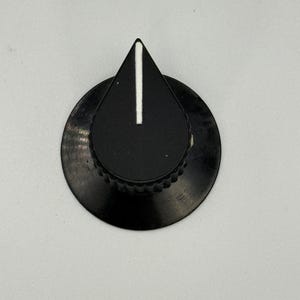 May include: A black control knob with a white vertical line indicator. The knob has a teardrop-shaped top and a circular base with a ridged edge. The knob is set against a plain white background.