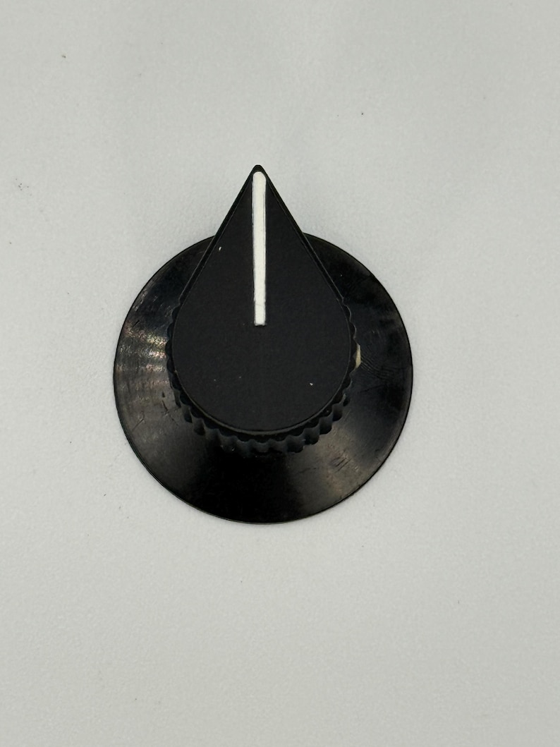 May include: A black control knob with a white vertical line indicator. The knob has a teardrop-shaped top and a circular base with a ridged edge. The knob is set against a plain white background.