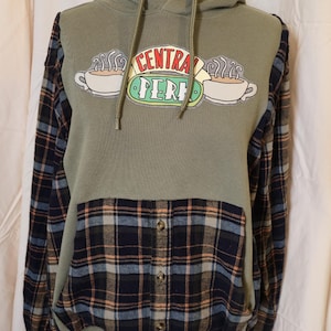May include: Olive green hoodie with a "Central Perk" logo and plaid sleeves and pocket. The hoodie has a drawstring hood and a buttoned pocket. The logo features two coffee cups and the text "Central Perk".
