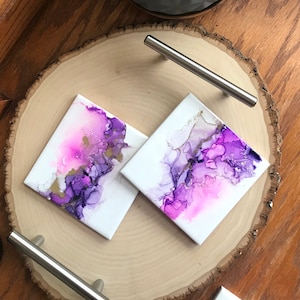 May include: Two square coasters featuring abstract purple and pink alcohol ink art. The coasters are displayed on a wooden slice with a silver handle.