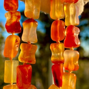 May include: Close-up of a beaded necklace featuring translucent, irregularly shaped beads in shades of orange, red, and yellow. The beads are strung together with a thin, light-colored string, creating a vibrant and eye-catching accessory.