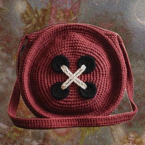 Puede incluir: Bolso de hombro de ganchillo burdeos con forma de botón grande. El bolso presenta cuatro acentos circulares negros y un diseño de punto de cruz blanco en el centro. La correa es del mismo color burdeos que el bolso.