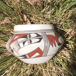 May include: A white ceramic pot with a geometric design in black and red. The pot has a rounded body and a slightly flared rim. The design features abstract shapes and lines. The pot is sitting in a bed of dry grass.