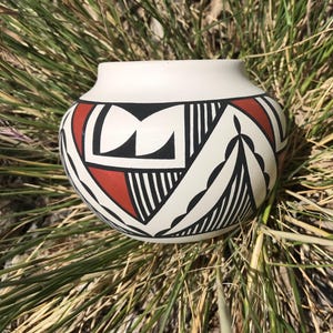 May include: A cream-colored ceramic pot with a rounded body and a wide opening. The pot is adorned with a geometric design featuring black, red, and white shapes. The design includes triangles, lines, and curved elements, creating a Southwestern aesthetic.