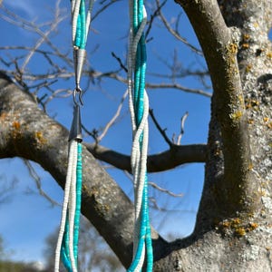May include: A multi-strand necklace with alternating strands of white and turquoise beads. The necklace is suspended from a tree branch against a clear blue sky. The clasp and connecting elements are silver-toned metal.