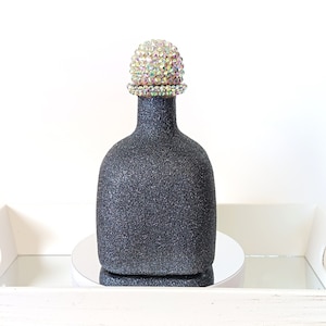 May include: A black bottle with a glittery finish and a crystal-encrusted stopper. The bottle is sitting on a white tray with a mirrored surface.