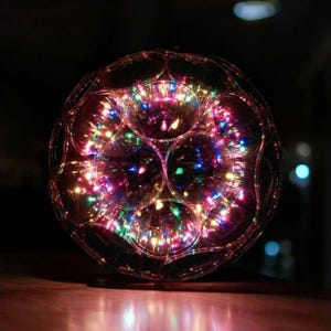 May include: A spherical decorative light fixture with a clear, segmented exterior. The interior is filled with numerous small, colorful LED lights, creating a vibrant display of red, green, blue, and yellow hues. The fixture sits on a wooden surface.
