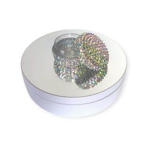 May include: A clear, round container with a lid encrusted with iridescent rhinestones. The container sits on a reflective, circular surface, creating a mirrored effect. The lid is partially open, revealing the inside of the container.