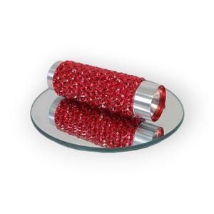 May include: A red, cylindrical object covered in small, sparkling crystals, with silver-colored metal ends, rests on a small, round mirror. The object's reflection is visible on the mirror's surface, creating a symmetrical image.