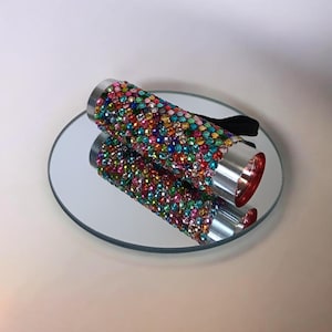 May include: A kaleidoscope with a cylindrical body covered in colorful rhinestones. The kaleidoscope has silver metal ends and a black strap. It is resting on a round, mirrored surface.
