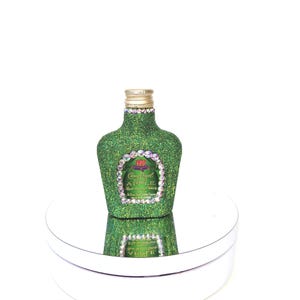 May include: A green glass bottle with a gold lid and a label that says "Crown Royal Whisky Apple" with a red crown logo. The bottle is covered in green glitter and rhinestones.