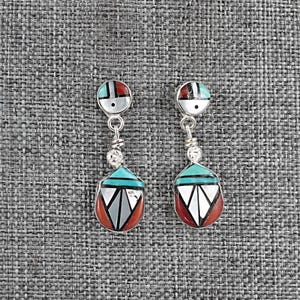 May include: A pair of silver earrings with geometric designs. Each earring features a circular top with red, turquoise, and white accents, connected to a teardrop-shaped pendant. The pendant has a turquoise top, white and gray geometric shapes, and a red bottom.