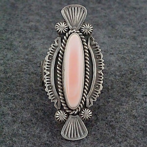 May include: A silver ring with a pink oval stone set in a detailed design. The ring has a rope-like border around the stone and intricate patterns on the sides.