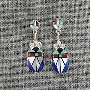 May include: A pair of silver earrings with inlaid geometric designs. Each earring features a small, round top with red, white, and turquoise accents, connected to a larger, teardrop-shaped pendant with a blue, black, red, and white pattern.