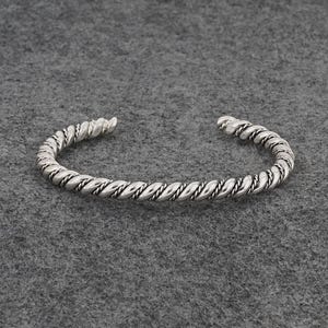 May include: Silver-colored cuff bracelet with a twisted rope design. The bracelet has an open design and is made of a shiny metal. The twisted pattern is consistent throughout the bracelet, creating a textured look.