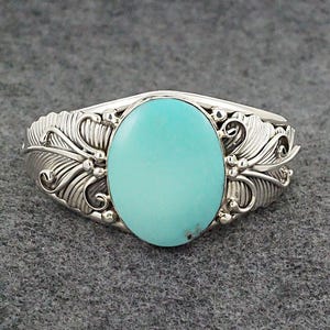 May include: A silver bracelet with a turquoise stone set in the center. The bracelet has a detailed design of feathers and swirls.