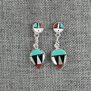 May include: A pair of silver-toned dangle earrings with a Southwestern design. Each earring features a round top with turquoise and red accents, connected to a teardrop-shaped pendant with black, white, and red geometric patterns. The earrings are displayed against a gray textured background.