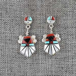 May include: A pair of silver-toned earrings with a Native American design. Each earring features a circular top with red, turquoise, and white accents, connected to a pendant with a geometric pattern in black, red, turquoise, and white.