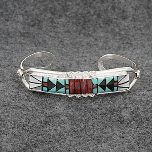 May include: A silver cuff bracelet with a rectangular centerpiece. The centerpiece features a mosaic design with turquoise, black, red, and white inlays. The bracelet has a decorative pattern along the edges.