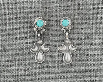 Puede incluir: Un par de pendientes plateados con una piedra turquesa en la parte superior y un diseño floral decorativo y estilizado. Los pendientes tienen forma de gota y se muestran sobre un fondo gris texturizado.