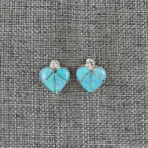 May include: A pair of heart-shaped earrings with a silver setting. The earrings feature a mosaic of light blue and turquoise-colored stones. The earrings are displayed on a gray textured surface.