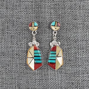 May include: A pair of silver-toned dangle earrings with geometric designs. Each earring features a circular top with red, blue, and gold accents, connected to a pendant with turquoise, red, and cream-colored inlays. The earrings have a southwestern style.