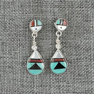 May include: A pair of silver-toned dangle earrings with a Southwestern design. Each earring features a circular top with red, turquoise, and white inlays, connected to a teardrop-shaped pendant with similar geometric patterns. The earrings are displayed against a gray textured background.