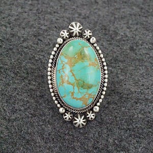 May include: A turquoise ring with a silver setting. The turquoise is a light blue color with brown veins. The setting is oval shaped and has a beaded edge. The ring is set with silver stars.