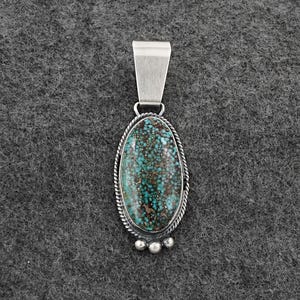 May include: Oval turquoise pendant with a silver bail and decorative silver rope trim. The turquoise stone has a mottled pattern of teal and brown. Three small silver beads are at the bottom of the pendant.