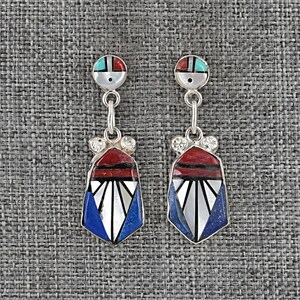 May include: A pair of silver earrings with a geometric design. Each earring features a circular top with turquoise and red accents, connected to a pendant with red, white, and blue inlays. The earrings are set against a gray textured background.