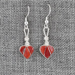 May include: A pair of heart-shaped earrings with red stone inlays. The earrings have a silver-colored metal setting and a hook closure. The earrings are displayed on a gray textured background.