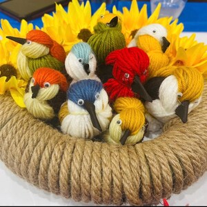 May include: A handcrafted bird nest wreath featuring colorful yarn birds with black beaks, nestled among yellow sunflowers. The wreath is constructed from coiled rope, creating a rustic and charming decorative piece.