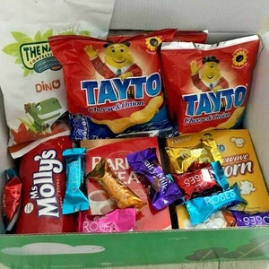 Può includere: Una scatola di cartone piena di vari snack. La scatola contiene sacchetti di patatine Tayto Cheese & Onion, snack Dino, caramelle Ms. Molly's, cioccolatini Cadbury Dairy Milk, cioccolatini Roses e popcorn per microonde.