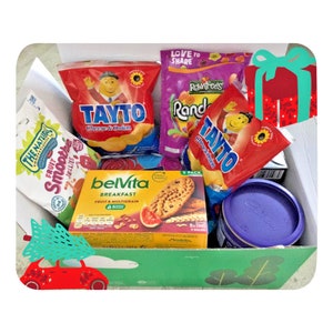 Op de afbeelding: Een groene en rode cadeaudoos gevuld met snacks. De doos heeft een kerstboom- en autodesign aan de voorkant. De snacks omvatten een zak Tayto kaas- en uienchips, een doos Belvita ontbijtkoekjes, een zak Rowntrees Randoms snoepjes en een zak Tayto kaas- en uienchips.
