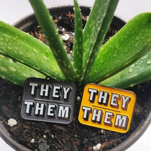 May include: Two enamel pins with the text "They Them" in white letters on a black and yellow background. The pins are sitting on top of soil in a pot with a green plant.