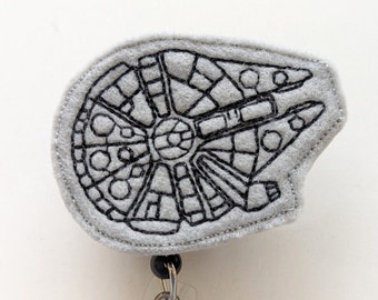 May include: A gray felt badge holder with a black embroidered image of the Millennium Falcon spaceship from Star Wars.